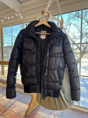 Svart pufferjacka från G-Star RAW - Snygg svart pufferjacka från G-Star RAW med krage, huva och flera fickor framtill. Jackan har bruna detaljer med logga på bröstet och ärmen. Perfekt för kalla dagar och har en modern, sportig look. Köpt för över 1000-lappen och är i ett fräscht skick. Priset kan diskuteras!