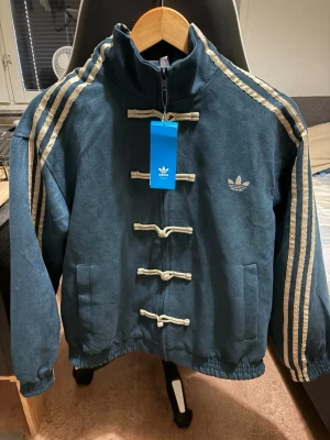 Adidas lunar/chinese jacka - Unik blå bomberjacka från adidas Originals med beige detaljer och snörning framtill. Jackan har klassiska tre ränder på ärmarna, hög krage och elastisk midja. Tillverkad i mjuk bomullsmix och har adidas logga broderad på bröstet. Väldigt unik jacka som är svårt att få tag på!