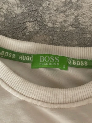 Vit långärmad tröja från Hugo Boss - Säljer en stilren vit långärmad tröja från Hugo Boss med rund halsringning och diskret logga på bröstet. Tröjan är i mjuk bomull och har ribbade muddar vid hals och ärmslut. Perfekt för dig som gillar klassisk och enkel stil.