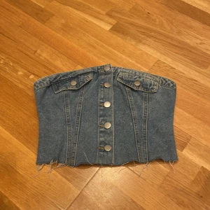 Blå jeans korsett topp med knappar - Säljer en snygg korsett topp i blå denim med silverfärgade knappar framtill och två dekorativa fickor. Toppen är axelbandslös och har råa kanter nedtill för en edgy look. Baksidan har smock för extra stretch och passform. 