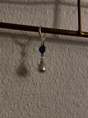 Blå och silver örhänge med pärla  - Säljer ett stilrent örhänge i silverfärg med en mörkblå sten och en droppformad vit pärla. Perfekt accessoar för att lyfta din look med en elegant touch. Passar till både vardag och mer uppklädda tillfällen.