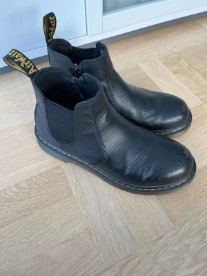 Svarta Dr. Martens chelsea boots - Klassiska svarta chelsea boots från Dr. Martens i skinn med elastiska paneler på sidorna och dragflikar med gul text. Grov, genomskinlig sula med gul söm runt om. Perfekta för dig som vill ha en tidlös och edgy stil.