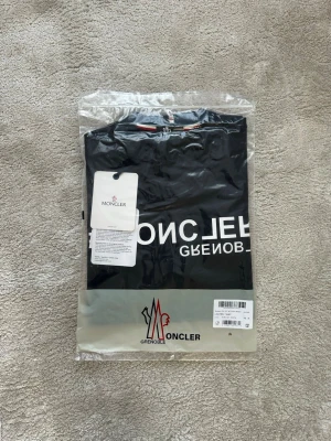 Svart Moncler Grenoble t-shirt - Svart t-shirt från Moncler Grenoble med vit printad logga och text framtill samt på ärmen. Klassisk rund halsringning och korta ärmar. Tillverkad i mjuk bomull för skön känsla. Perfekt för dig som vill ha en clean och stilren look med street vibe.