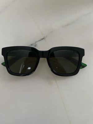 Svart-grön Gucci solglasögon - Säljer ett par feta solglasögon från Gucci med svart front och gröna skalmar med röd rand. Klassisk fyrkantig form och tydlig Gucci-logga på sidorna. Modell GG0001SN. Perfekt accessoar för dig som vill sticka ut.