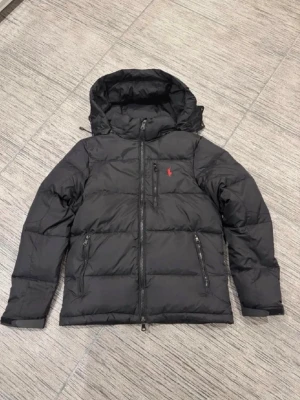 Svart pufferjacka från Polo Ralph Lauren - Säljer en svart pufferjacka från Polo Ralph Lauren med röd broderad logga på bröstet. Jackan har huva, tre dragkedjeförsedda fickor och en rak passform. Perfekt för kalla dagar och riktigt snygg!