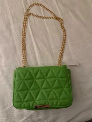 Grön axelväska- Glitter - Snygg grön axelväska från Glitter och guldfärgad kedjerem. Väskan har en stilren rektangulär form och detaljer i guldmetall. Perfekt för att lyfta vilken outfit som helst med en pop av färg.