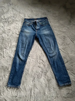 Steady Eddie II ”Mid Worn” Nudie  - Säljer ett par klassiska blå jeans från Nudie Jeans Co med snygga slitningar och orangea sömmar. Modellen har fem fickor och en cool detalj med broderad bakficka. Jeansen är raka i benen och har normal passform. Perfekt för dig som gillar tidlös stil. Storlek 29x32
