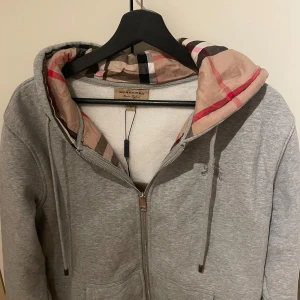 Grå hoodie från Burberry - En fin kofta från Burberry i bra skick, en vardaglig kofta som också kan användas till finare tillställningar. Koftan är väldigt bekväm och samtidigt stilren. Storlek L men passar även bra för folk som bär storlek M