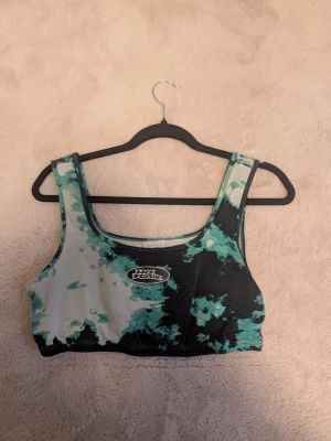 No Fear camo linnetopp i grönt och svart - Säljer en sportig linnetopp från No Fear med grönt, svart och vitt camouflage-mönster. Toppen har breda axelband och rundad halsringning, perfekt för träning eller chill. Materialet är mjukt och stretchigt polyester som sitter skönt mot kroppen.