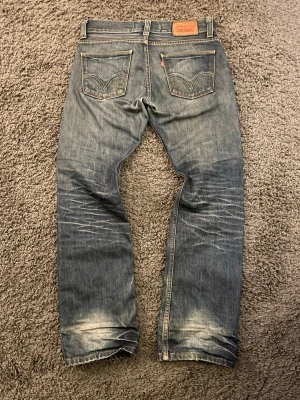 Levi's 506 jeans i blå denim - Säljer ett par klassiska Levi's 506 jeans i blå denim med snyggt slitna detaljer och kontrastsömmar. Jeansen har raka ben, fem fickor och läderpatch bak i midjan. Perfekta för dig som gillar en tidlös och avslappnad look. Osäker på storlek men midjan är 41cm, längd 106 benöppning 20.