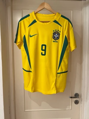 Brazil home kit 2002 X Ronaldo 9 - Säljer en officiell Brasilien fotbollströja från Nike med nummer 9 och Ronaldo på ryggen. Tröjan är gul med gröna detaljer och har Brasil-logga på bröstet. Perfekt för dig som älskar fotboll och vill ha en ikonisk landslagströja.