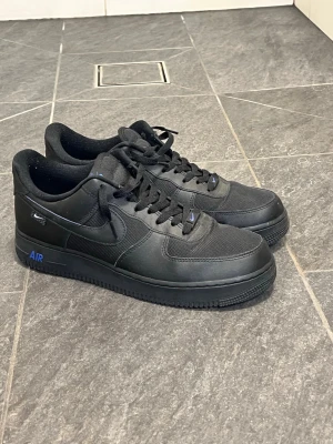 Nike Air Force 1 svart/blå sneakers - Säljer ett par svarta Nike Air Force 1 sneakers med blå detaljer och klassisk siluett. Skorna har ovandel i skinn och mesh, svart sula och blå Nike-logga på plösen och innersulan. Perfekta för dig som vill ha en clean och tidlös look. I stort sett helt nya och bara använda ett fåtal gånger. Hör av er vid frågor.