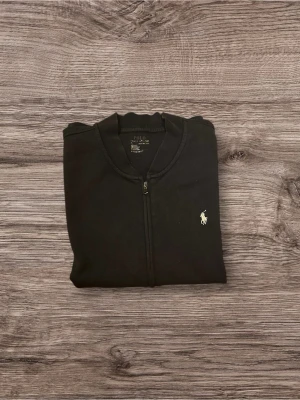 Mörkblå Zip tröja från Polo Ralph Lauren - Klassisk mörkblå zip tröja från Polo Ralph Lauren med dragkedja framtill och diskret broderad logga på bröstet. Tröjan har rund halsringning, långa ärmar och ribbade muddar. Perfekt för dig som gillar stilrena och tidlösa plagg.