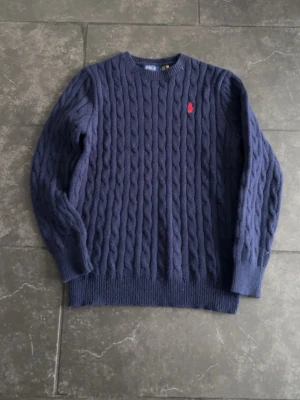Mörkblå stickad tröja Polo Ralph Lauren - Säljer en mörkblå kabelstickad tröja från Polo Ralph Lauren i storlek S. Tröjan har ett klassiskt mönster med grova flätor och en röd broderad logga på bröstet. Rund hals och ribbade muddar vid ärmslut och nederkant. Perfekt för kyliga dagar!