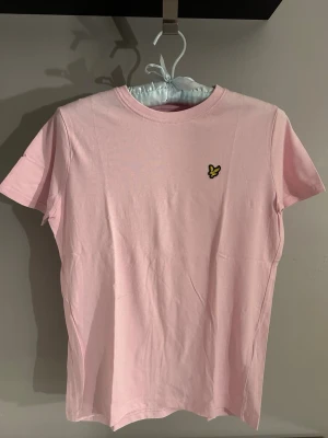 Ljusrosa t-shirt från Lyle & Scott - Säljer en ljusrosa t-shirt från Lyle & Scott Junior med klassisk rund hals och korta ärmar. T-shirten har den ikoniska gula örnloggan broderad på bröstet och är gjord i 100% mjuk bomull. Perfekt för en clean och stilren look.