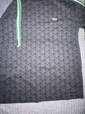 Svart mönstrad t-shirt från Lacoste - Snygg svart t-shirt från Lacoste med diskret geometriskt mönster över hela tröjan. Klassisk rund halsringning och gröna samt vita ränder på axlarna. Ikoniska krokodilloggan på bröstet. Perfekt för dig som gillar streetwear och vill ha något unikt.