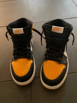 Nike Air Jordan 1 svart/gul sneaker - Säljer ett par Nike Air Jordan 1 sneakers i svart, vitt och gul-orange. Klassisk high-top modell med svart swoosh, vita paneler och gula detaljer på tå, häl och sula. Skorna har snörning och är tillverkade i skinn för en clean och sportig look.