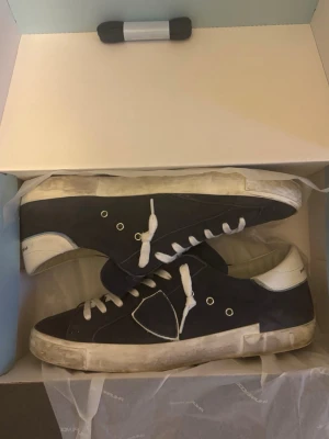 Philippe Model blå sneakers - Snygga sneakers från Philippe Model i mörkblå mocka med vita detaljer på hälen och vita snören. Skorna har en rund tå, platt sula och klassisk logga på sidan. Perfekta för dig som gillar stilrena och sportiga skor med en fräsch vibe. Låda och sko påse kommer med.