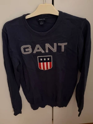 Mörkblå GANT tröja med tryck - Snygg mörkblå tröja från GANT i storlek M med stort broderat GANT-tryck och amerikanskinspirerat emblem på bröstet. Tröjan har rund hals och långa ärmar, tillverkad i mjuk bomull som är skön mot huden. Perfekt för dig som gillar klassisk och sportig stil.