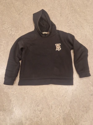 Burberry hoodie - Snygg och stilren Burberry hoodie i svart med den ikoniska TB-monogramloggan och Burberry-text tryckt på ryggen. Klassisk design som passar perfekt både till vardags och för en mer uppklädd streetwear-look.