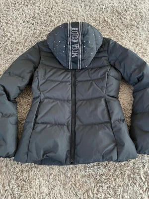 Svart dunjacka från Moon Boot med Swarovski kristaller - Säljer en svart pufferjacka i dun från Moon Boot med Swarovski kristaller på huvan och logga . Jackan har dragkedja framtill, lång ärm och quiltad design. Perfekt för kalla dagar när du vill sticka ut lite extra. Jackan är helt ny och oanvänd. Storlek 174 cl, vilket är som en Small. 