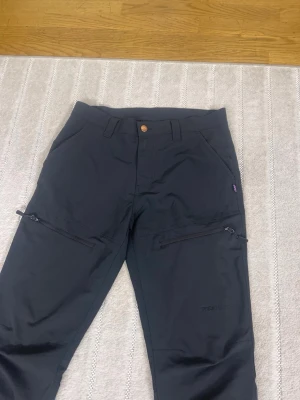 Svarta tekniska täckbyxor med dragkedjor - Säljer ett par svarta tekniska täckbyxor från Tenson. Byxorna har raka ben, flera fickor med dragkedjor och bälteshällor. Materialet är slitstarkt och passar perfekt för utomhusaktiviteter. Snygg och clean look med diskret logga på benet.