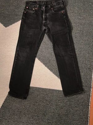 Levi's 501 svarta jeans W30 L30 - Säljer ett par klassiska svarta Levi's 501 jeans med raka ben och knappgylf. Jeansen har fem fickor, silverfärgade knappar och nitar samt den ikoniska läderlappen bak i midjan. Tillverkade i slitstark bomullsdenim med en snygg tvättad finish.