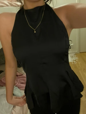 Svart omlott blus med hög hals - Säljer en svart blus med hög hals och omlott-detalj i midjan. Toppen är ärmlös och har en lätt glansig finish som ger en lyxig känsla. Passformen är normal och materialet känns mjukt och följsamt mot huden. Perfekt att styla med smycken eller jeans.