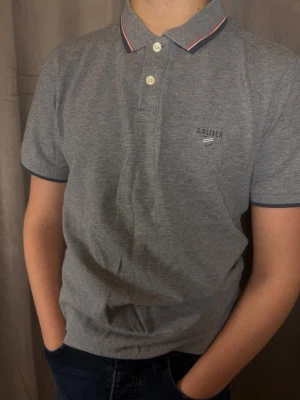 Grå pikétröja från S.Oliver - Säljer en stilren grå pikétröja från S.Oliver med smal passform. Tröjan har korta ärmar, två knappar framtill och en krage med marinblå och röda detaljer. Materialet är mjukt och skönt, perfekt till vardags. S.Oliver-logga broderad på bröstet.