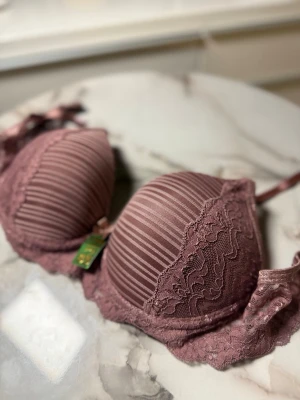 Randig spets-bh i dammrosa - Supersnygg bh i dammrosa med tunna ränder och detaljer i spets. Kuporna är vadderade och har en liten rosett framtill. Sidorna är dekorerade med genomskinlig spets som ger en romantisk vibe. Perfekt för dig som gillar feminina detaljer.