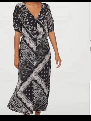 Svart långklänning med vit mönster - Supersnygg långklänning från H&M i svart med vitt paisley- och patchworkmönster. Klänningen har v-ringning, korta puffärmar och en avslappnad passform. Perfekt för dig som gillar boho-vibbar och vill ha något unikt i garderoben.
