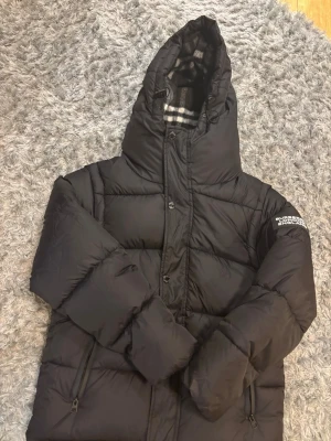 Svart pufferjacka från Burberry - Säljer en svart pufferjacka från Burberry med klassiskt rutigt foder i huvan och tydlig logga på ärmen. Jackan har dragkedja, knappar och två fickor med dragkedja. Perfekt för kalla dagar och riktigt snygg till streetwear-stilen.