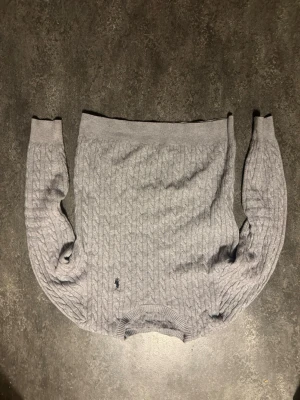 Grå kabelstickad tröja från Polo Ralph Lauren - Säljer en grå kabelstickad tröja från Polo Ralph Lauren med klassisk svart logga broderad på bröstet. Tröjan har rund hals, ribbade muddar och lång ärm. Perfekt för lager på lager under höst och vinter.