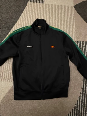 Svart track jacket från Ellesse - Säljer en svart track jacket från Ellesse med gröna och gula ränder längs ärmarna. Tröjan har hel dragkedja, hög krage och två fickor framtill. Klassisk logga på bröstet och sportig vibe. Perfekt till streetwear-stilen.