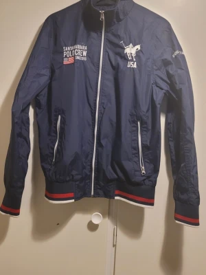 Marinblå vindjacka från Vinson Polo Club - Säljer en marinblå vindjacka från Vinson Polo Club med vita och röda detaljer. Jackan har dragkedja framtill, två fickor med dragkedja och broderade loggor på bröstet och ärmen. Ribbstickade muddar med röd och vit rand ger en sportig look.