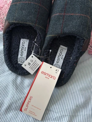 Rutiga tofflor från Esmara Men stl 41 - Mysiga herrtofflor från Esmara i storlek 41. Utsidan har ett mörkgrått rutigt mönster med röda och gula linjer, och insidan är fodrad med mjuk, mörk textil för extra komfort. Sulan är svart och räfflad för bra grepp.