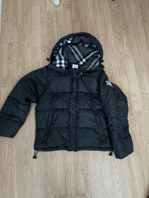Svart pufferjacka från Burberry - Säljer en svart pufferjacka från Burberry med klassiskt rutigt foder i huvan. Armarna går att ta av för att göra den till en Väst, Storlek S
