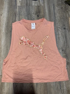 Rosa linnetopp från Puma Kenza - Säljer en ljusrosa linnetopp från Puma x Kenza med ett stort blommigt Puma-tryck framtill. Toppen är ärmlös och har en croppad passform, perfekt för sommardagar. Tillverkad i mjuk bomull som känns skön mot huden.