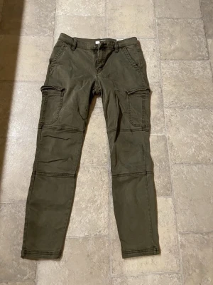 Olivgröna cargopants från H&M L.O.G.G. - Säljer ett par olivgröna cargopants från H&M L.O.G.G. med flera fickor på benen och bakficka med lock. Byxorna har normal passform och är tillverkade i mjuk bomull. Perfekta för en avslappnad och cool stil.