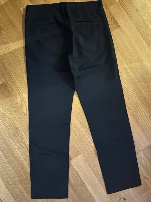 Svarta kostymbyxor från Boomerang - Snygga svarta kostymbyxor med raka ben och klassisk passform. Byxorna har två bakfickor, varav en med knapp, samt hällor för bälte. Materialet känns mjukt och stilrent, perfekt till en dressad look. Passar dig som gillar enkel och clean stil.