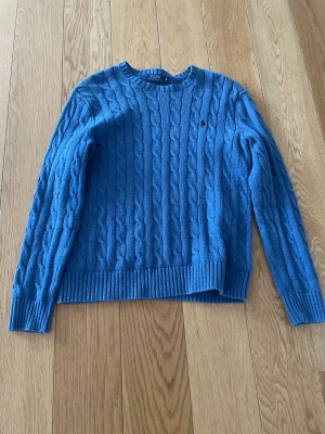 Blå kabelstickad tröja Polo Ralph Lauren - Kabelstickad blå tröja från Polo Ralph Lauren i storlek S. Klassisk rund halsringning och ribbade muddar vid ärmslut och nederkant. Perfekt för dig som gillar en tidlös och stilren look.