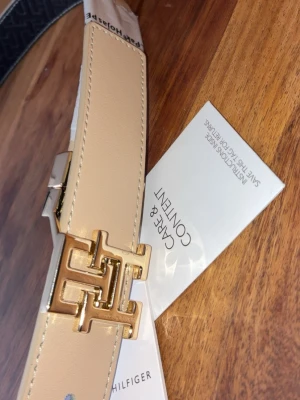 Beige och svart | Reversibel bälte | Tommy Hilfiger - Snyggt vändbart bälte från Tommy Hilfiger i beige och svart med guldfärgat TH-spänne. Ena sidan är slät beige och andra sidan är svart med präglat TH-mönster. Perfekt accessoar för att lyfta vilken outfit som helst. 🚨ALDRIG ANVÄNT