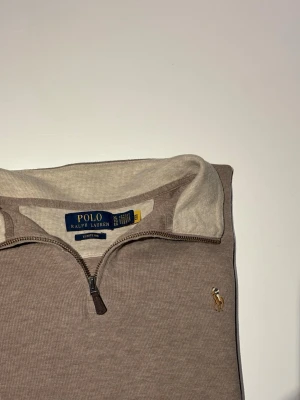 Ralph Lauren Half Zip - Elegant half zip-tröja från Polo Ralph Lauren, tillverkad i den exklusiva Estate Rib-kvaliteten som ger en mjuk, följsam och lyxig känsla. Tröjan har en tidlös design i en stilren beige/ljusbrun nyans, med klassisk halv dragkedja och den ikoniska broderade Polo-logotypen på bröstet.  Perfekt för både vardag och mer uppklädda tillfällen – lika snygg över en skjorta som till ett par chinos eller jeans.  Nypris: 2195 SEK