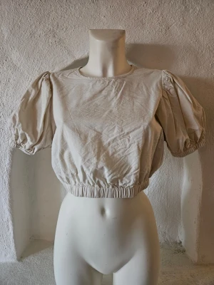 Beige croppad blus med puffärm - Säljer en beige croppad blus med rund hals och puffiga korta ärmar. Toppen har resår vid midjan och ärmarna vilket ger en snygg siluett. Tillverkad i mjuk bomull som känns skön mot huden. Perfekt för dig som gillar stilrena och trendiga plagg.