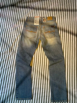 Nudie Jeans Grim Tim Silver Indigo W31 L30 - Säljer ett par Nudie Jeans Grim Tim i tvätten Silver Indigo. Klassiska blå jeans med raka ben, orange sömmar och bakfickor med Nudie-signatur. Tillverkade av 100% ekologisk bomull för en schysst känsla och look. Helt oanvända!