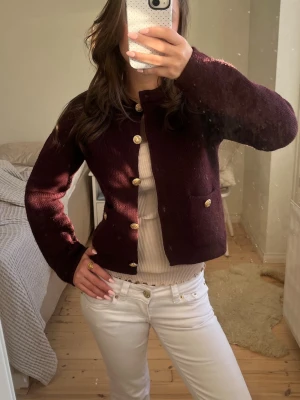 Vinröd kofta med guldknappar - Vinröd kofta med raka, långa ärmar och snygga guldknappar framtill samt på fickorna. Koftan är stickad och har en klassisk, lite boxig passform. Perfekt att slänga över en topp för en chic look.