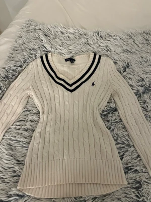 Vit kabelstickad tröja Ralph Lauren - Kabelstickad vit tröja från Ralph Lauren med marinblå broderad logga på bröstet. Djup v-ringning med två marinblå ränder runt halsen. Långärmad och ribbade muddar. Perfekt för en preppy look!
