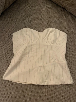 Vit randig korsett från Bershka - Säljer en snygg vit korsett från Bershka med tunna svarta ränder. Toppen har bandeau-modell och snörning i ryggen för justerbar passform. Materialet känns som bomullsmix och ger en stilren och trendig look. Perfekt att styla med jeans eller kjol.