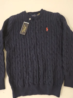 Mörkblå stickad tröja Polo Ralph Lauren - Kabelstickad mörkblå tröja från Polo Ralph Lauren med klassisk röd broderad logga på bröstet. Tröjan är i 100% bomull, storlek M men passar som S. 