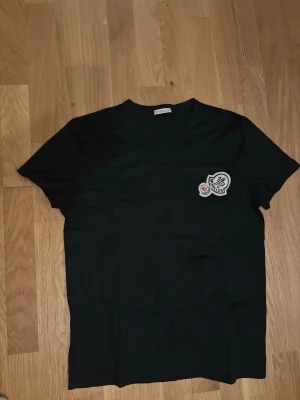 Svart Moncler t-shirt med patch - Snygg svart t-shirt från Moncler med klassisk rund hals och korta ärmar. På bröstet sitter två broderade Moncler-patchar i vitt, blått och rött som ger en clean men exklusiv vibe. Tillverkad i mjuk bomull för skön känsla hela dagen.
